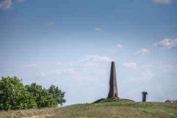 Stone obelisk on the crest of Zagajicka brda in Vojvodina, Serbia, stands alone against a wide blue sky, surrounded by grassy slopes and sparse trees, conveying the remoteness of the Banat sand hills.