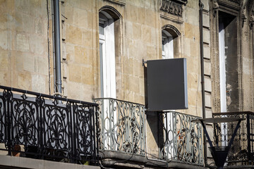 Empty blank grey square sign fixed between two tall white windows, ready for future commercial branding on a sunny day on a french building.