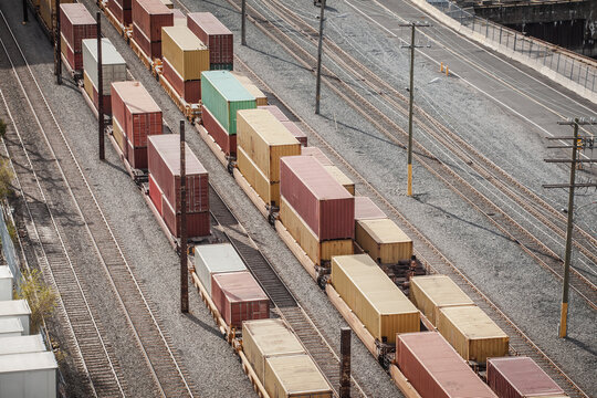 Rows of double stacked freight containers sit on flatbed railcars in a busy port railyard, parallel tracks converging toward an industrial pier that handles North American import export traffic.