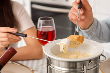 Young couple with glass of wine dipping croutons into cheese fondue at home, closeup