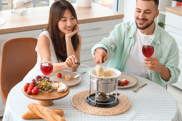 Young happy couple with glasses of wine dipping croutons into cheese fondue at home