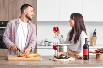 Young happy couple with glasses of wine cutting bread for dipping in cheese fondue at home