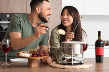 Young happy couple with glasses of wine dipping croutons into cheese fondue at home