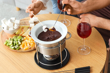 Young couple with glasses of wine dipping sweet snacks into chocolate fondue at home, closeup