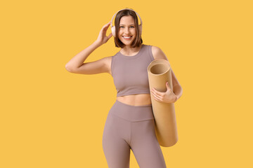Sporty young woman in headphones with fitness mat on yellow background