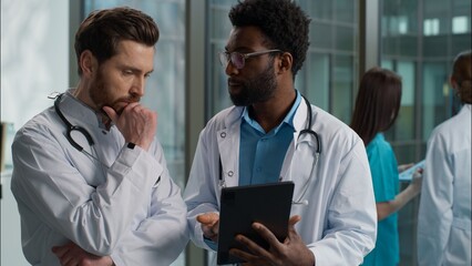 Multiethnic doctors conversation African American therapist and Caucasian surgeon physician men talking brainstorming diagnosis holding tablet device thinking difficult case at hospital clinic hallway