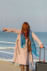 Ginger-haired woman, 30s, standing on pier with suitcase. Happy and stylish in coat and scarf....