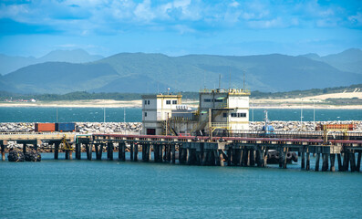 industrial pier with buildings, water, and distant mountains under a blue sky, depicting a working...