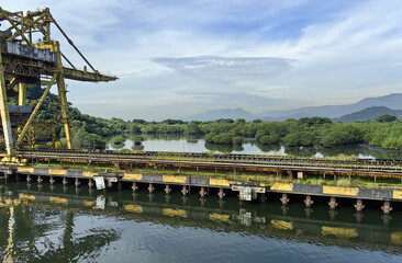 Overgrown industrial pier with rusting structures beside a calm river and dense green forest, with...