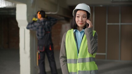 Happy multinational builders Asian woman engineer mobile phone call communication smartphone construction coordination safety helmet planning Caucasian man tools drilling noise distraction renovation