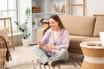 Beautiful young woman sitting on floor and using mobile phone in living room