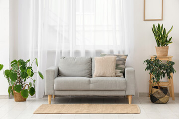 Interior of light living room with grey sofa, stool and houseplants near big window