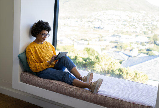 African American woman in sweater sitting on home bench by large window reading tablet, copy space