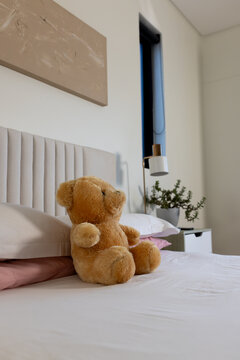 Teddy bear is sitting upright on bed with white sheets, pink pillows, abstract art, near nightstand