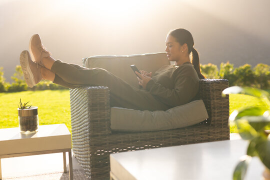 Woman lounging in wicker chair and scrolling phone, sun backlighting lawn, table with succulent
