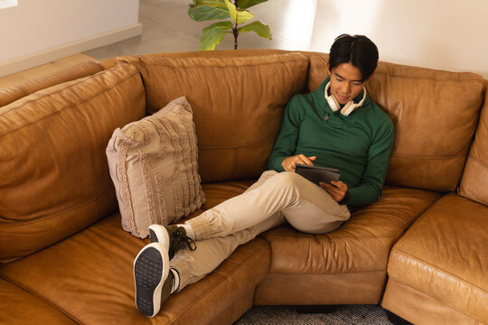 Asian man sitting on leather couch in living room using tablet with headphones and throw pillow