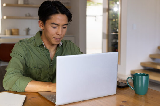 Asian man working on silver laptop at kitchen dining table while using green mug and smartphone