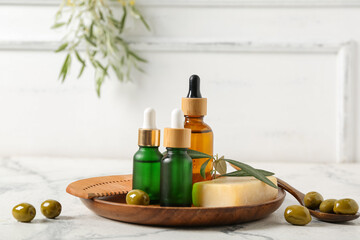 Bottles of essential olive oil, soap bar and wooden comb on marble table near white wall, closeup