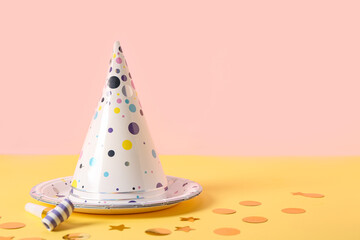 Composition with party hats, whistle and paper plate on yellow table near pink wall, closeup