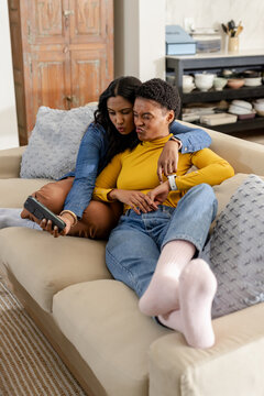 Diverse female couple leaning together on beige sofa in living room puckering lips at smartphone