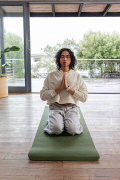 Non-binary person wearing smartwatch kneeling and praying on green yoga mat by sliding glass doors