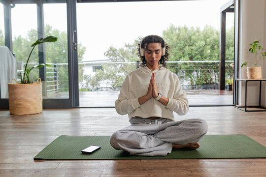 Non-binary adult meditating on green yoga mat at home, with smartphone beside, wearing headphones