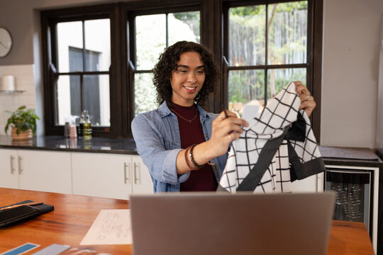 Non-binary person holding checkered top over laptop at kitchen counter with sketches, swatches
