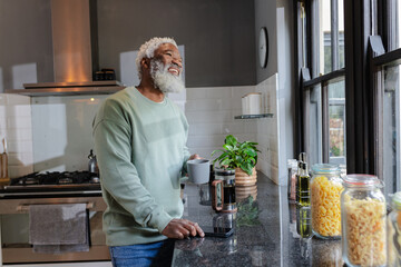 African American Senior man holding gray mug and gazing out window in kitchen with French press