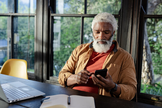 Senior African American man checking smartphone at home office desk with laptop, notebook and pen