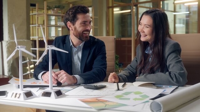 Happy joy Caucasian businessman Korean businesswoman discuss wind turbine model pointing finger laugh talk eco energy office startup sustainable development meeting cooperation innovation paper office