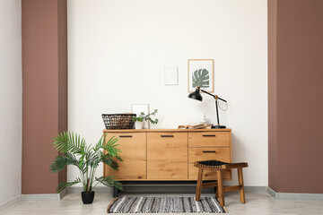 Interior of living room with chest of drawers, stool and houseplant near white wall