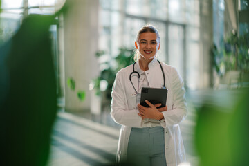 Female doctor smiling and holding tablet in hospital corridor