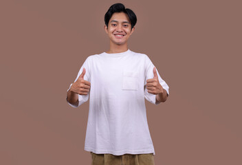 Happy Young Asian Man in White T-shirt Shows Thumbs Up Over Brown Background