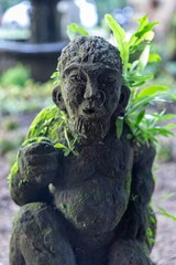 A moss-covered stone statue of a person stands in a garden in Ubud, Bali, Indonesia. The statue is partially covered in moss and small plants, showing nature reclaiming art.