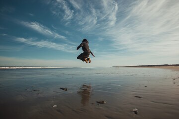 A person jumping in the air on a beach with a striped shirt and brown pants under a cloudy blue sky