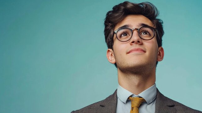 A young man with curly hair and glasses, wearing a suit and tie, looking up against a blue background.
