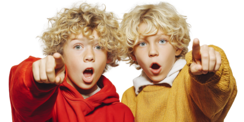 Two excited children with curly blonde hair, wearing colorful sweaters, express surprise and joy while pointing at something off camera