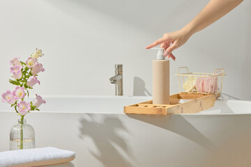 Elegant bath corner with hand reaching for beige bottle on ledge. Natural light from window casts...