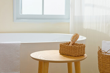 Soft daylight enters a minimalist bathroom with round wood table beside tub. A brush and basket...
