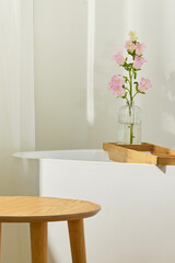 Soft bathroom light with white tub, curtain and wooden tray. Delicate flowers add warmth. The tray...