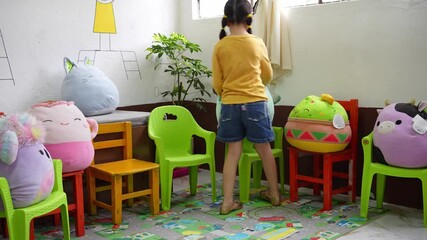 kindergarten girl placing varied stuffed toys arranged on classroom chairs as if attending a lesson in a preschool. Imaginative preschool classroom setup, and early childhood playful learning
