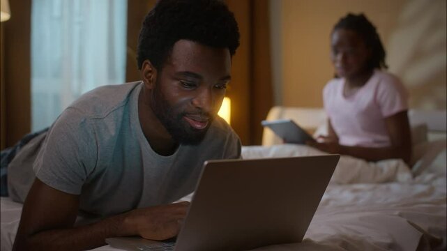 African American family husband using laptop wife digital tablet evening bedroom ethnic couple relaxing lying bed weekend gadget addiction scrolling social media watching device separation home night - Powered by Adobe