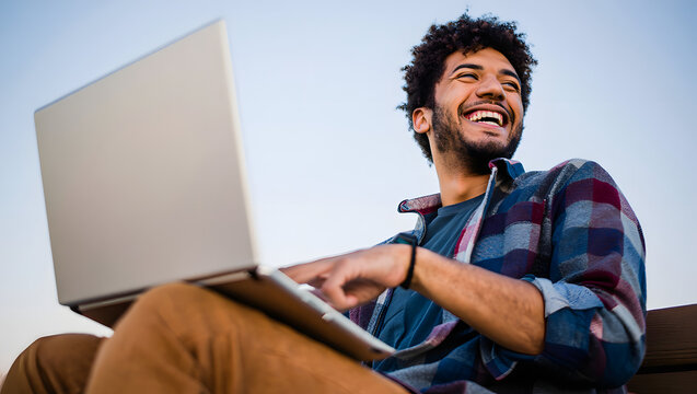 Young man with curly hair laughing while working on a laptop outdoors