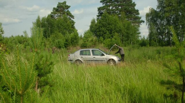 Automotive trouble plagues driver as he examines his malfunctioning car engine, stranded amidst field of tall grass, trees, and cloudy skies. Frustrated driver inspecting broken car engine.