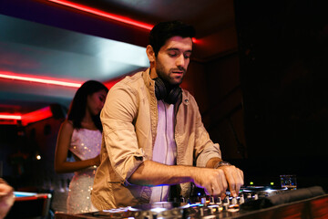 Male DJ focused on mixing music at nightclub, wearing headphones and casual jacket. Vibrant red...