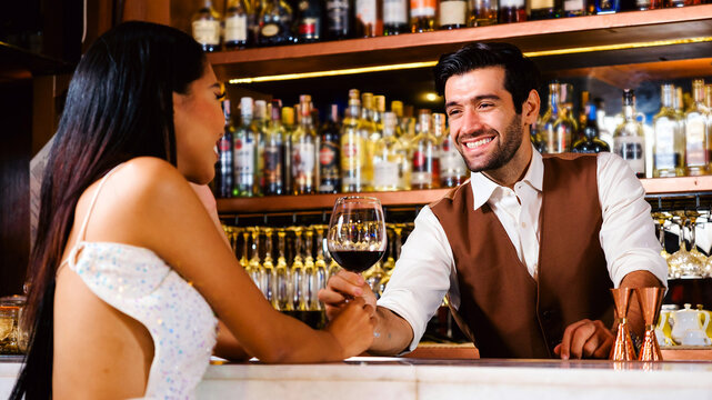 Smiling bartender chats with a stylish woman holding a wine glass at an elegant bar. Warm lighting and shelves of bottles set a friendly, upscale nightlife scene with relaxed, social vibes.