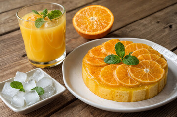 shows a wooden table with a plate of orange-flavored cake topped with slices of orange and mint leaves, a glass of orange juice with a mint leaf, and a small dish of ice cubes with mint leaves.