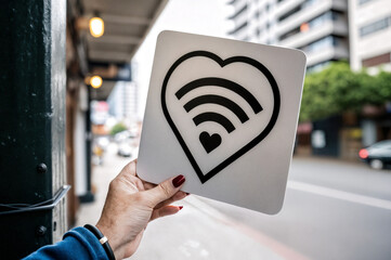 a hand is holding a white square sign with a black heart shape containing a Wi-Fi signal icon, indicating a love for wireless technology or a place offering free Wi-Fi. The background shows an urban s