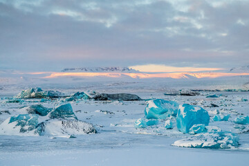 Icebergs and huge chunks for green-blueish ice fill Jokulsarlon Glacier lagoon, as the golden hour sunlight of the early morning casts a beautiful warm glow over the glacier-filled national park in