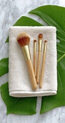 Set of four makeup brushes, light beige/tan handles with gold accents, on a cream-colored towel, atop a marble surface with tropical leaves